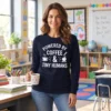 Woman wearing a Powered by Coffee and Tiny Humans long sleeve shirt, holding a coffee mug in a classroom.