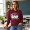 Woman wearing an “I’m the Cool Teacher (According to Me)” long sleeve shirt beside a classroom desk.